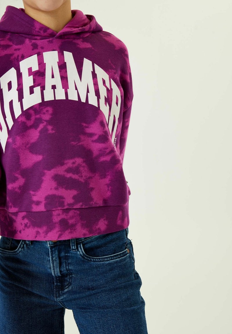Personne portant un sweat à capuche tie-dye violet avec le mot « DREAMER » en grandes lettres blanches et un jean bleu sur un fond clair uni.