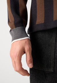 Primo piano di una mano appoggiata accanto a un corpo che indossa un maglione a strisce marroni e nere con polsini grigi e jeans di denim neri.