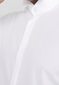 Camicia bianca con colletto a punta, chiusura con un solo bottone e tessuto liscio e strutturato. Il design è classico e aderente.