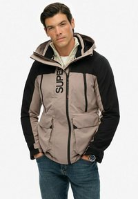 Grå og sort zip-up jakke med flere lommer, hætte og logo detalje. Den har en glat tekstur og et sporty design.