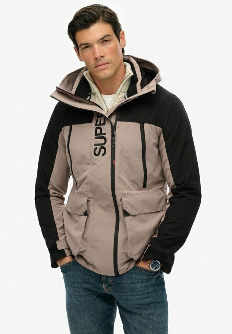 Grå og sort zip-up jakke med flere lommer, hætte og logo detalje. Den har en glat tekstur og et sporty design.