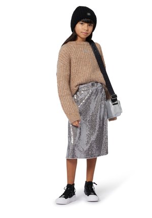 Chunky beige gebreide trui, midi-jurk in zilveren pailletten, zwarte beanie en zwarte sneakers met witte zolen. Kleine zilveren crossbody tas.