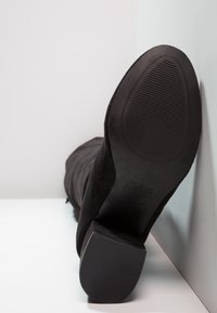 Botín negro de ante hasta el tobillo con una suela texturizada. Presenta un tacón grueso y un zipper lateral. Marca mínima visible en la suela.