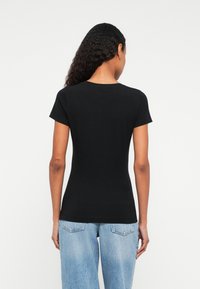 Kvinne med krøllete hår står med ryggen til, iført en stram svart kortermet skjorte og lyseblå høytlivsjeans.