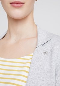 Blazer grigio con una texture liscia, dotato di una spilla decorativa. Sotto, una maglietta bianca con righe orizzontali gialle.