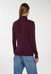 Burgundowy sweter z dzianiny z wysokim kołnierzem i ściągaczem. Długie rękawy z dekoracyjnymi guzikami. Połączony z jasnoniebieskimi, wysokimi spodniami dżinsowymi.