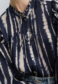 Blusa a righe navy e crema con colletto arricciato, chiusura frontale con bottoni e cravatta al collo. Presenta strisce verticali e tessuto testurizzato.