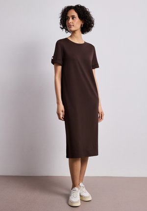 Vrouw met krullend haar die een knielange bruine jurk draagt met opgerolde korte mouwen en witte sneakers, staand tegen een effen grijze achtergrond.