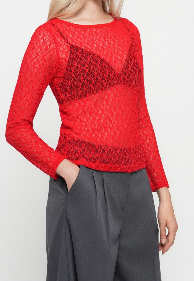 Femme portant un haut en dentelle rouge à manches longues sur un bralette noir, associé à un pantalon ample gris taille haute, une main dans la poche.