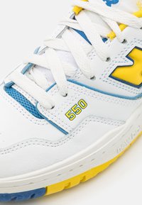 Zapatilla New Balance 550 blanca con acentos en azul y amarillo, cordones blancos y el número amarillo "550" en el panel lateral.