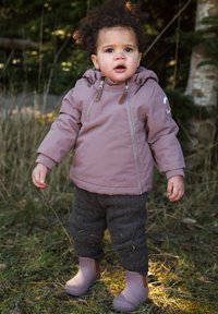 Veste imperméable rose avec accents marron clair, pantalon en laine gris et bottes en caoutchouc roses. Enfant debout sur l'herbe avec un feuillage vert en arrière-plan.