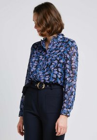 Blouse transparente marine avec motif floral en violet et bleu, dotée d'un col et de manches longues, associée à un pantalon sombre taille haute.