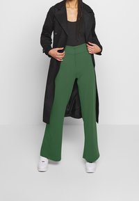 Persoon in groene wijde broek, zwart tanktop, lange zwarte jas en witte sneakers, staand tegen een effen lichte achtergrond.
