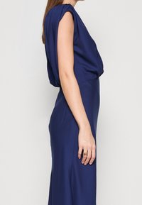 Mörkblå klänning med draperad design, korta puffärmar och en figursydd silhuett. Slät tygtextur, visad från sidan.