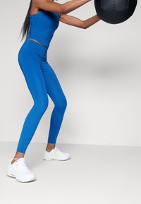 Top corto atletico blu e leggings in un tessuto liscio e elastico. Sneakers bianche con accenti al neon. Persona impegnata in un allenamento.