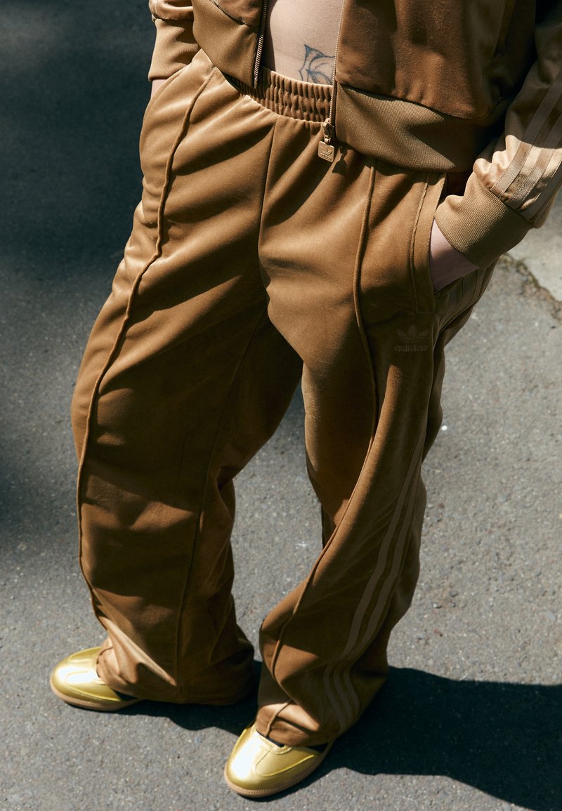Brune velour joggebukser med sidelister, elastisk midje og frontlommer, kombinert med skinande gullsko på en teksturert overflate.