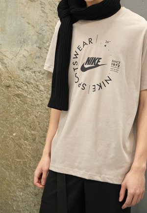 Osoba ubrana w beżową koszulkę Nike Sportswear z czarnym tekstem i logo oraz czarny szalik zarzucony na jedno ramię.