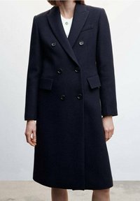 Cappotto doppiopetto in lana blu navy con rever a lancia, due tasche frontali e bottoni neri. Texture liscia con un design lungo fino al ginocchio.