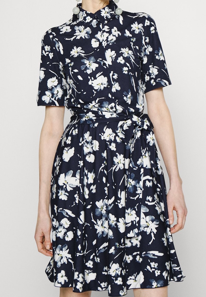 Femme portant une robe bleu marine avec imprimé floral blanc, manches courtes, col et ceinture nouée à la taille.