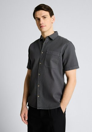 Młody mężczyzna stojący z jedną ręką w kieszeni, ubrany w ciemnoszarą, teksturowaną koszulę na guziki z krótkim rękawem na jednolitym tle.