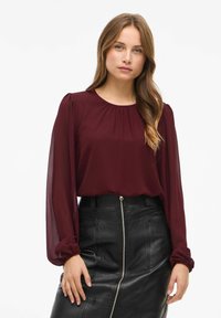 Blusa bordeaux con scollatura rotonda, maniche lunghe trasparenti e dettagli arricciati. Abbinata a una gonna a tubino in pelle nera con zip frontale.