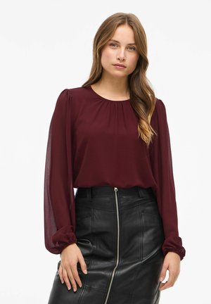 Blusa burdeos con escote redondo, mangas largas transparentes y detalle fruncido. Combinada con una falda lápiz de cuero negra con cremallera frontal.