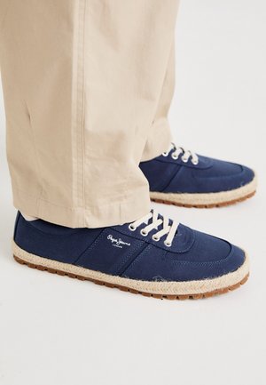 Chaussures décontractées bleu marine avec lacets blancs et bordure en corde, portées avec un pantalon beige sur fond blanc.