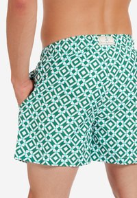 Costume da bagno verde realizzato in tessuto leggero, con un motivo geometrico bianco, una cintura elastica e tasche laterali.