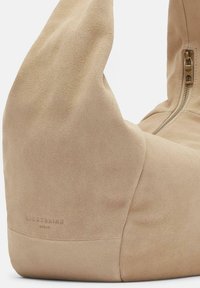 Sac en suède beige avec une forme pliée unique, doté d'une fermeture éclair latérale et d'un logo de marque en relief pour plus de détails. Texture douce sur l'ensemble.
