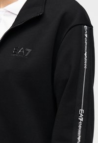 Zwarte sweatshirt met een kraag met rits. Heeft een reliëflogo op de borst en een verticale streep met bedrukt tekst langs de mouw.