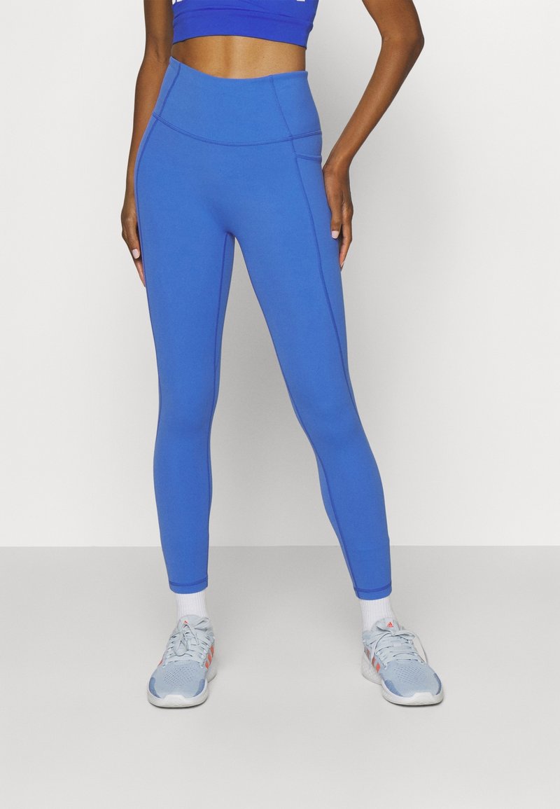 Modré legíny s vysokým pasem vyrobené z hladkého, elastického materiálu s plochými švy a těsným střihem, doplněné o světle modré sportovní boty.