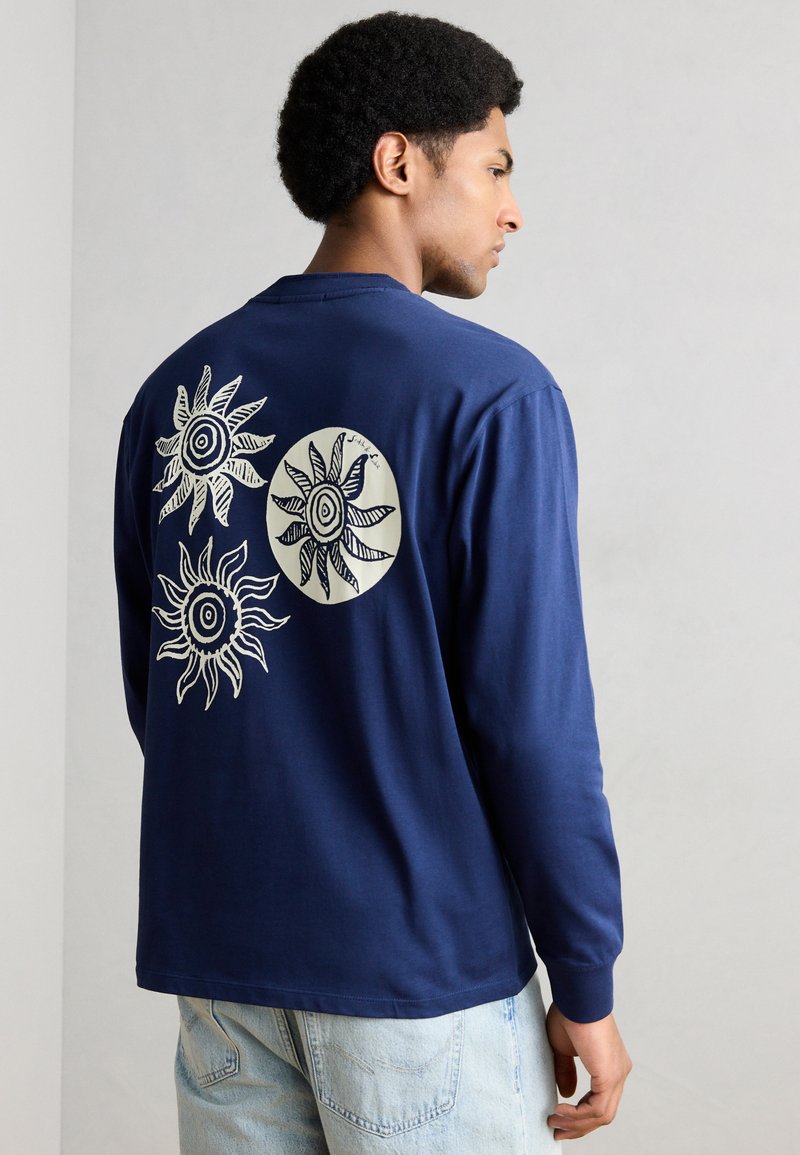 Μπλε μπλούζα με μακριά μανίκια από βαμβάκι, με τρία λευκά σχέδια ήλιων στην πλάτη, με ένα κυκλικό μοτίβο ήλιου ως κεντρικό στοιχείο.