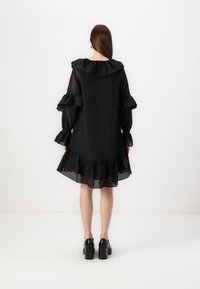 Svart, volangkjole med teksturert stoff og lagdelt kant. Lange, puffede ermer har volangdetaljer. Båret med svarte platåsko.