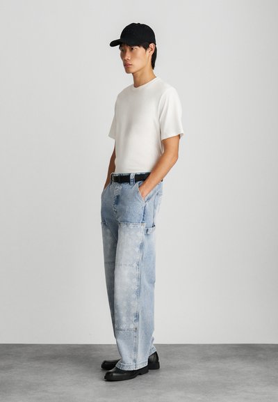 Camiseta blanca de manga corta combinada con jeans de mezclilla holgados de color azul claro. Cinturón negro y zapatos negros de punta redonda y lisa. Diseño simple, ajuste casual.