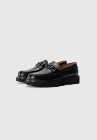Black leather loafers with a smooth finish, round toe, and decorative metal hardware. Thick black rubber sole with a textured pattern.