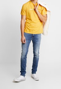 Žluté bavlněné tričko s logem "TOMMY", v kombinaci s světle modrými denimovými džíny a bílými teniskami. Jedinec drží krémovou bundu přes rameno.