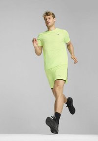 Neon zielona koszulka sportowa i dopasowane szorty z czarnymi akcentami. W granatowych butach do biegania, w dynamicznej pozycji biegowej na neutralnym tle.