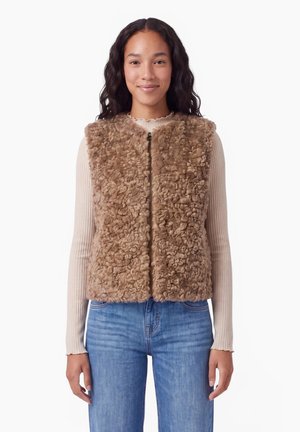 Mujer joven que lleva un chaleco de piel sintética rizado de color marrón claro sobre una camiseta beige de manga larga con textura acanalada y vaqueros azules, de pie sobre un fondo blanco.