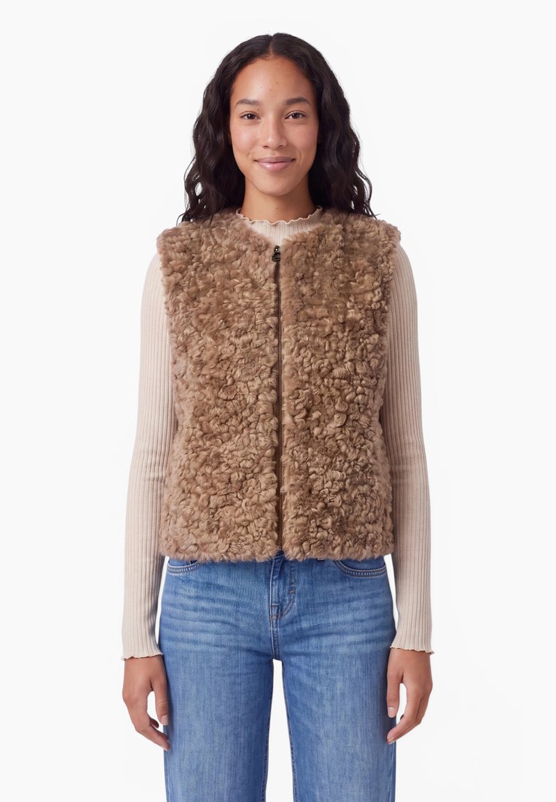 Jeune femme portant une veste sans manches en fausse fourrure bouclée marron clair sur un haut côtelé beige à manches longues et un jean bleu, debout devant un fond blanc.