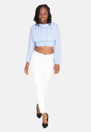 Vrouw met gevlochten haar draagt een lichtblauw cropped blouse met lange mouwen, witte aansluitende broek, zwarte hakken en een lichtbeige enkelarmband.