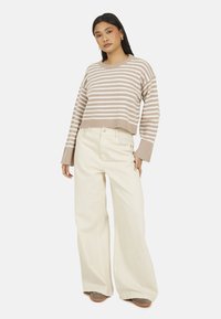 Donna che indossa un maglione a maniche lunghe beige e bianco a righe, pantaloni beige a gamba larga e scarpe marroni, in piedi con una mano in tasca.