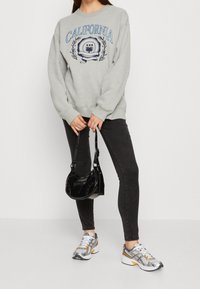 Person som bär en grå "California" sweatshirt, svarta jeans, silver- och orange sneakers, håller en liten svart handväska med båda händerna.