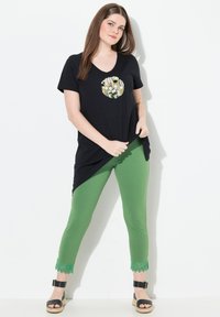 Camiseta negra de corte en V con un patrón floral circular, combinada con leggings verdes con detalles de encaje en el dobladillo y sandalias negras.