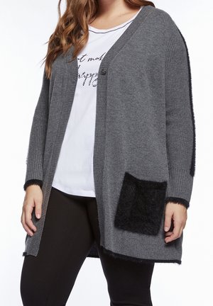 Femme portant un long cardigan gris avec une poche noire duveteuse et une bordure, par-dessus un t-shirt blanc imprimé et un pantalon noir, posant les mains détendues.