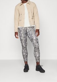 Giacca beige con bottoni sopra una t-shirt bianca, abbinata a leggings grigi mimetici aderenti e scarpe da atletica nere. Tessuto liscio, vestibilità su misura.