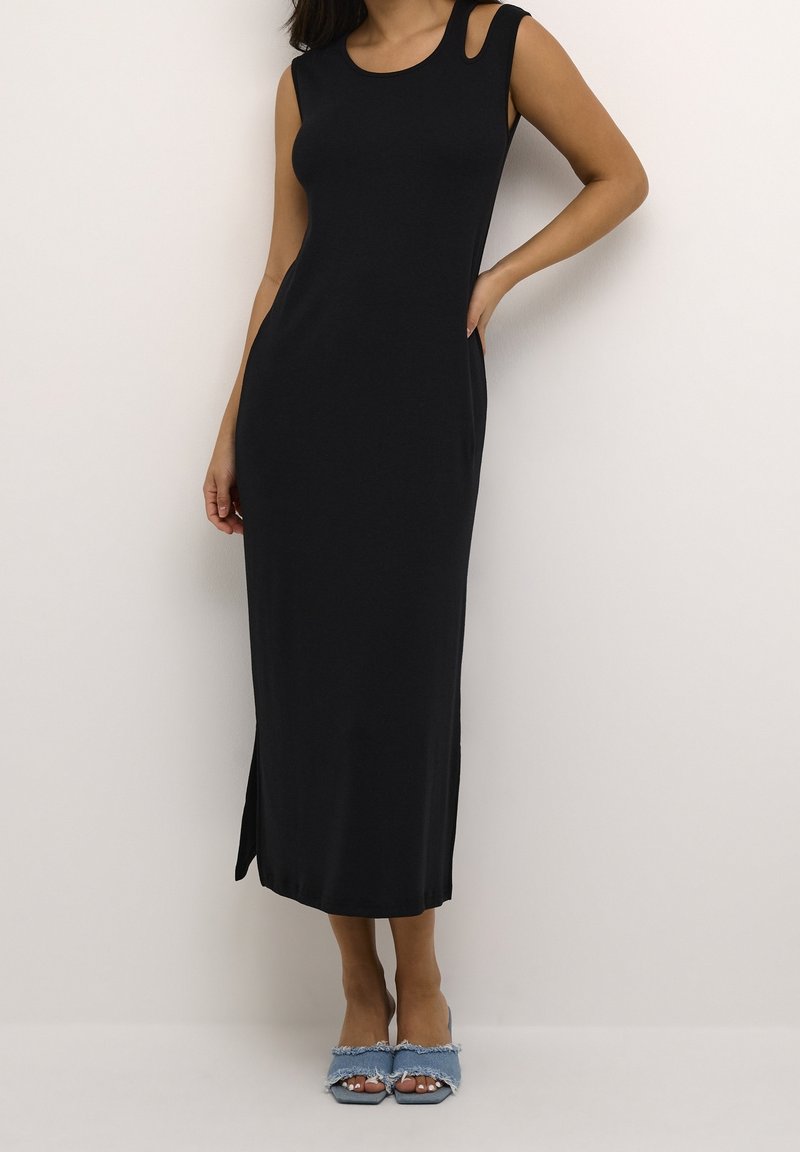 Femme portant une robe midi noire sans manches avec une découpe à l'épaule, debout avec une main sur la hanche et portant des talons ouverts en denim.