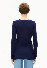 Langsleeve marineblauwe top van zacht materiaal, met een ronde halslijn en zijsplitten aan de zoom. Model wordt van achteren getoond.