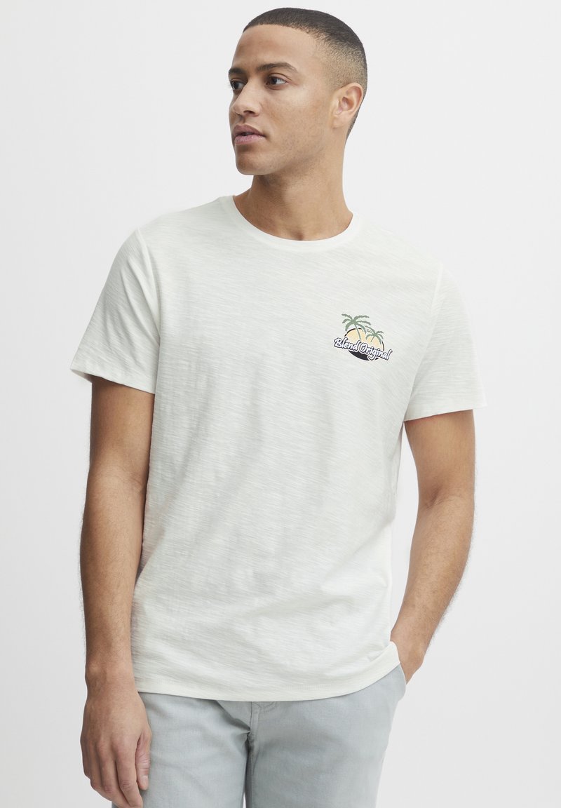 Lysegrå kortermet t-skjorte laget av et teksturert stoff, med et lite brodert design av to palmer og "Blend Original" på brystet.