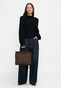 Czarny sweter w prążki i ciemne dżinsy o szerokich nogawkach. Modelka trzyma brązową skórzaną torbę na ramię z dwoma uchwytami i minimalistycznym designem.