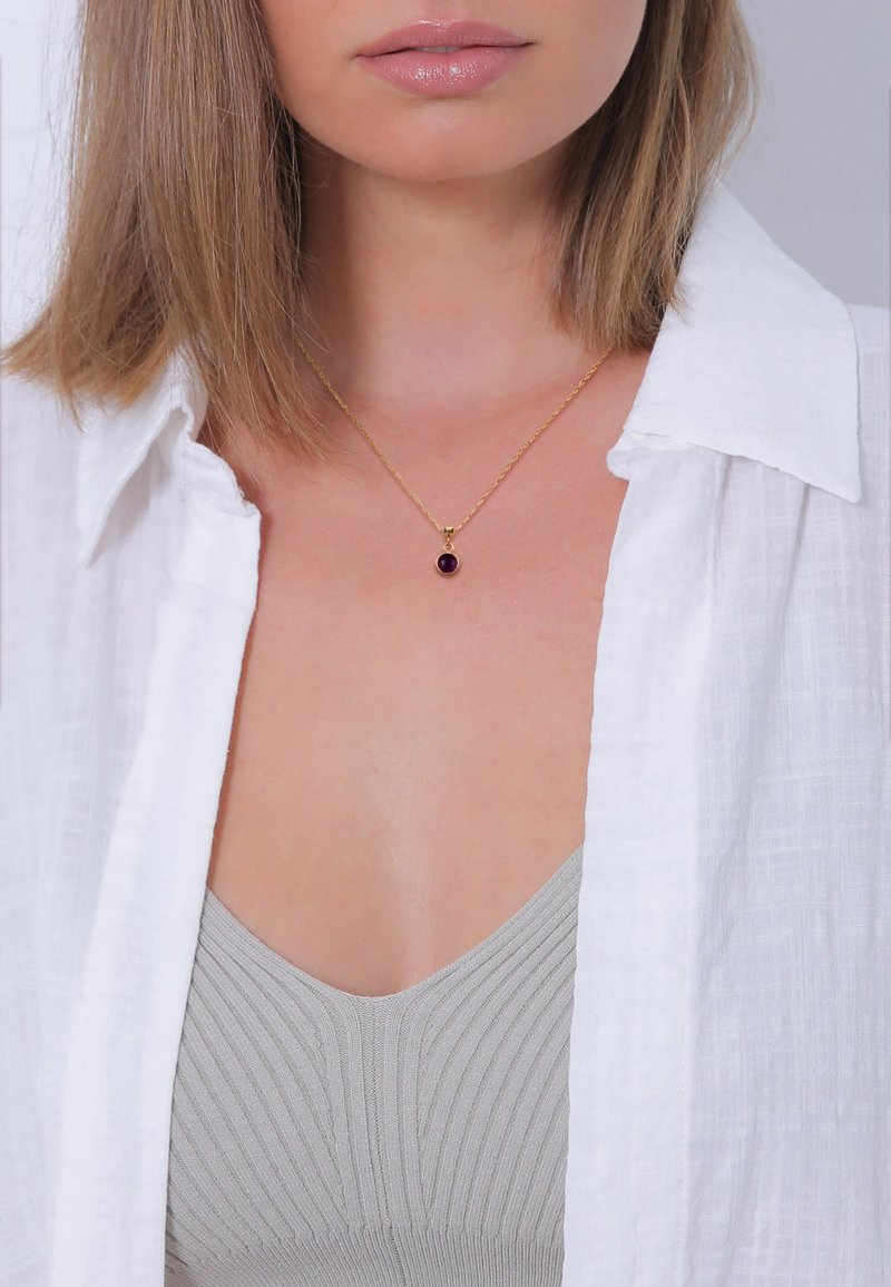Guldhalskæde med en dyb lilla oval vedhæng, båret over en lysegrå ribbet top, suppleret med en hvid struktureret skjorte med knapper.